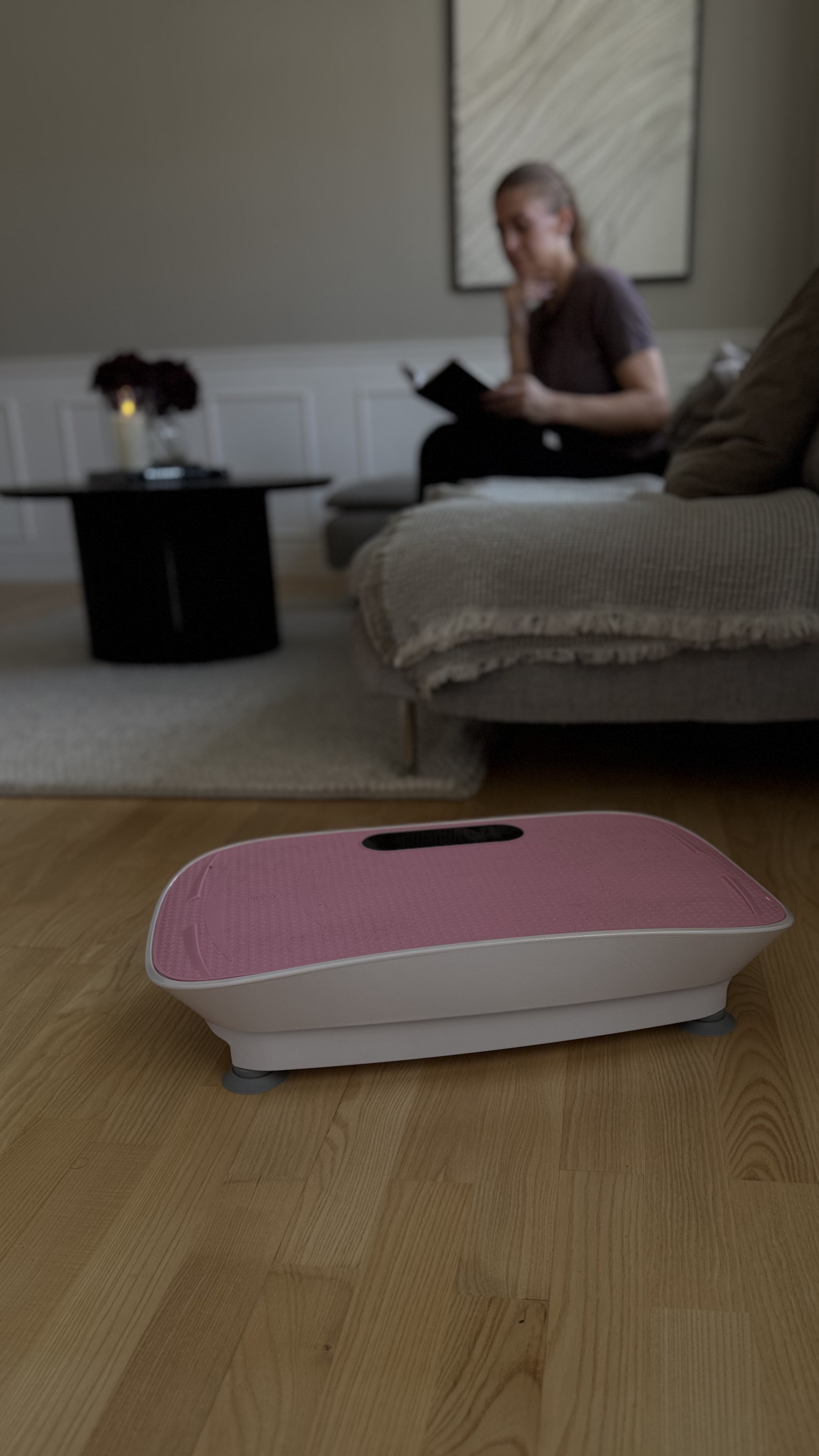 Pink vibration plate on a wooden floor with Debbie reading on a sofa in the background
